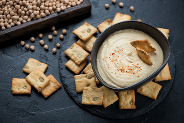 Bowl of hummus and crackers with pumpkin seeds, studio shot over black stone surface, elevated view