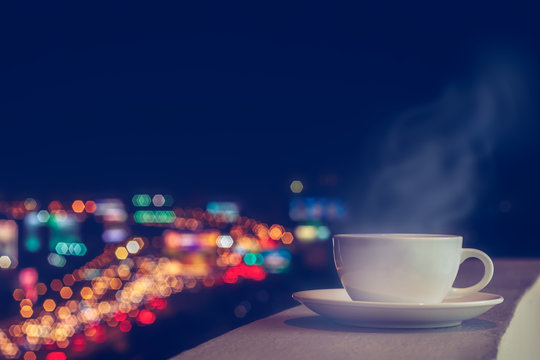White coffee cup and softy stream smoke put on sky terrace border with blurred twilight high angle view of city scape and colorful traffic lighting on road. Selective focus and copy space