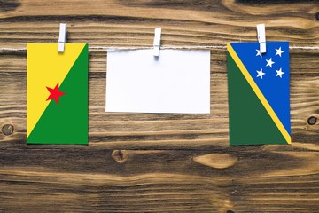 Hanging flags of French Guiana and Solomon Islands attached to rope with clothes pins with copy space on white note paper on wooden background.Diplomatic relations between countries.