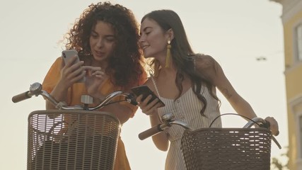 Beautiful young girls friends speaking and watching something on their smartphones while sitting on their bicycles outdoors at the street - Powered by Adobe