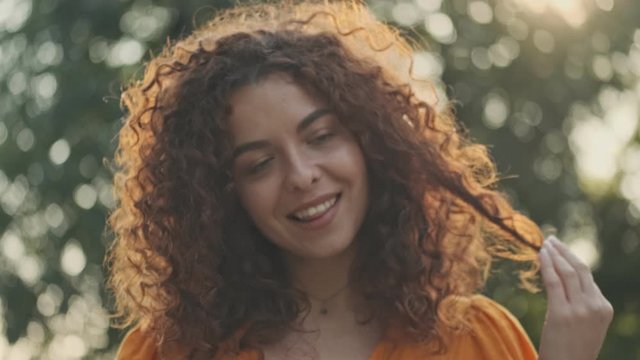 Beautiful young redhead woman in orange dress smiling and playing with her curly hair while resting in the park