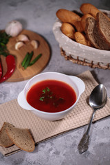 Tasty and hearty dinner. A plate with borsch on the table, next to the board is parsley, dill, green onions, garlic, chili pepper and a basket with bread.