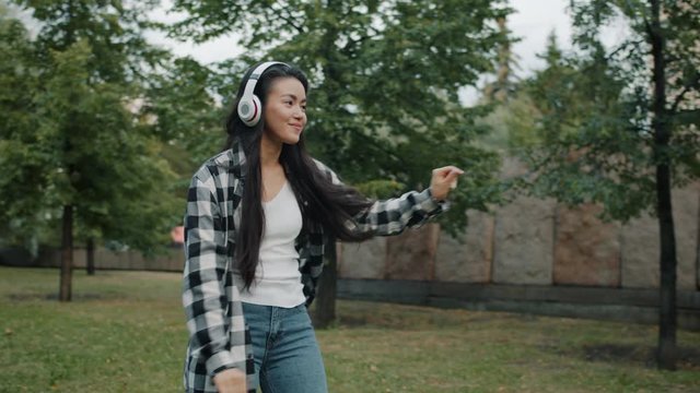 Slow motion of Asian lady dancing outdoors in city park wearing headphones having fun outdoors on summer day. Youth and modern culture concept.