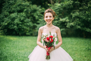 summer wedding photo: a young slim brown-haired bride girl stands in the background of a green park with a beautiful wedding bouquet of bright red color 