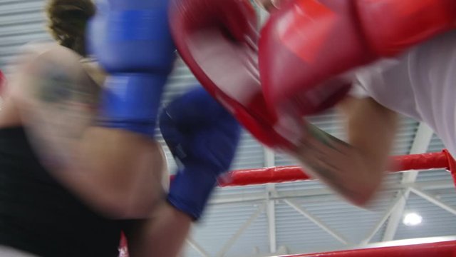 Low angle shot of professional female fighter in gloves hitting her partner as she holding hands in defensive position in corner of boxing ring during training