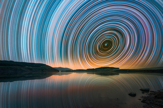 Beautiful night landscape with small lake and mountains. The colorful star trails on the sky. Night time lapse photography..
