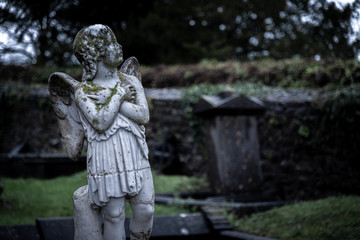 KILKENNY, IRELAND, DECEMBER 23, 2018: Sculpture of an old creepy cherub angel in the middle of a graveyard, full of lichen and mold, holding its chest and heart while looking at the cloudy sky.