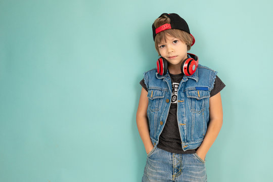A cool tenager boy in denim jaket and shorts, red earphones, black cap, stands in front of the camera and keeps his hands in pockets, isolated on blue background