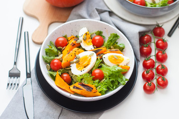 Lunch bowl with cherry tomatoes, roasted pumpkin, green salad, seeds and boiled eggs. Grey plate on white background. 