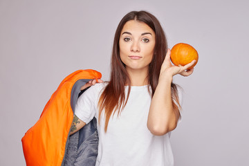 Shot of happy smiling lady has happy mood, carries small orange pumpkin, ready to cook vegetable dish from organic product, wears white shirt and bomber on shoulder, isolated on grey. Autumn concept.