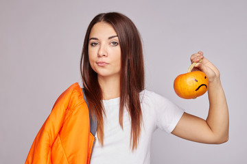 Horizontal shot of indecisive puzzled female with straight shiny hair, looks with hesitant expression aside, holds little pumpkin, poses against grey concrete wall with copy space advertisement, text.