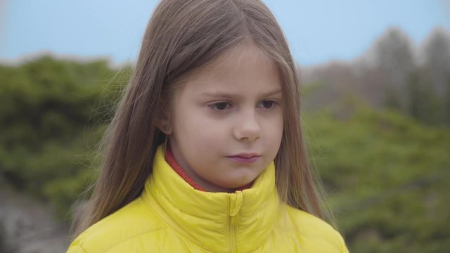 Close-up face of a pretty Caucasian girl with brown eyes listening to her mother or sister and shaking head. Cute child dressed in yellow coat standing in the autumn park.