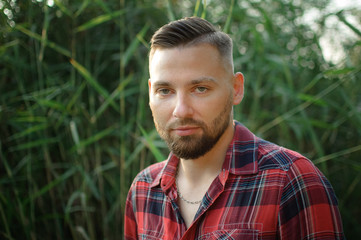 Brutal bearded young man in red shirt is standing outdoors on green stems background. Hipster style concept.