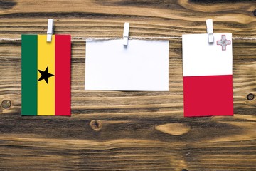 Hanging flags of Ghana and Malta attached to rope with clothes pins with copy space on white note paper on wooden background.Diplomatic relations between countries.