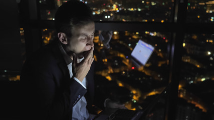 Sleepy young man in suit is working on his computer at night near the window with panoramic city view. He is taking off his glasses and rubbing tired eyes. Overtime work concept.