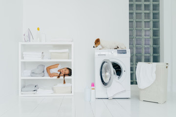 Little tired girl did work about house to help parents, sleeps on shelf near folded linen, iron and washing powder containers, jack russell terrier on washing machine. Children, house chores concept