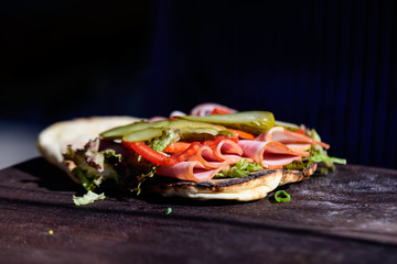 One fresh healthy Brazilian sandwich or burger with ham, green salad and pickled cucumbers is being prepared on a brown wooden table at a street food market festival, side with soft focus