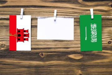 Hanging flags of Gibraltar and Saudi Arabia attached to rope with clothes pins with copy space on white note paper on wooden background.Diplomatic relations between countries.
