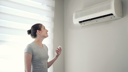 Young brunette woman in her modern apartment. She turns on the air conditioner from the remote...