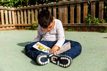 Fototapeta premium Niño con pluri discapacidad leyendo, mirando, jugando con un libro de colores en el suelo de un parque de ciudad.