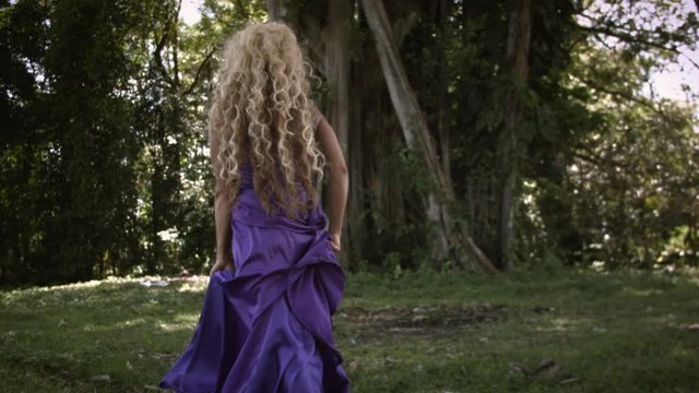 Young beautiful woman with curly blonde hair in elegant purple dress walking and posing on meadow near big tree with green foliage in tropical forest.