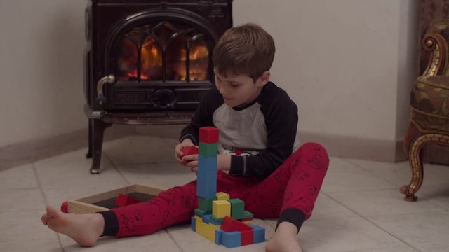 Autistic boy in christmas pajama sitting on the floor by the fireplace and playing with wooden blocks in slow motion. 