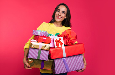 A bunch of happiness. Attractive dark-haired woman is looking happily at the camera, holding a great amount of multicolored present boxes in her hands.