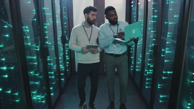 Two server engineers inspecting computer security rack corridor at data center. Multi-race IT technicians talking monitoring super computers in server rack cabinets.
