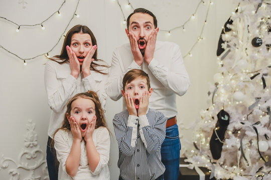 Image of young parents and their kids on new year background, with mouthes widely opened and hands on cheeks, looking at the camera with horror in eyes.