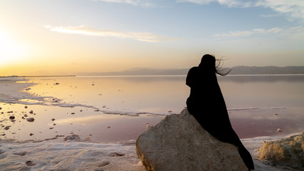 Unidentified Iranian woman in hijab burka sitting on a rock by the Maharloo pink lake, Shiraz, Iran
