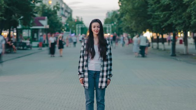 Zoom-in time lapse of beautiful young woman hipster standing outdoors in urban street smiling looking at camera while crowds of people are moving around.