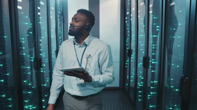 African IT administrator holding tablet walking in server rack corridor inspecting computer working performance at big data center room.