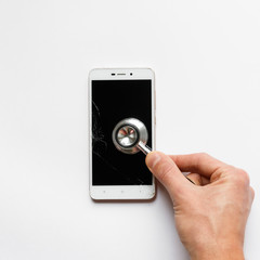Closeup of man hand holding stethoscope on broken mobile smartphone after drop, view from above, on white background. Copy space. Smart phone with cracked screen or touchscreen and damaged case.