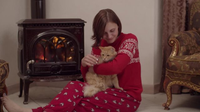 Young female in red christmas pajamas sitting on the floor with red cat on her legs by the fireplace and taking picture on her smartphone in slow motion 
