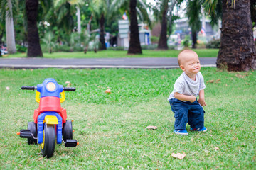 Cute little Asian 1 year old toddler baby boy child standing and playing near his tricycle in summer park, kid playing toy and cycling in the garden outdoors on nature Y