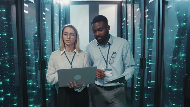 Female IT administrator talking with african server technician cooperating inspecting rack server computer cabinets in data center or cyber security. Teamwork.