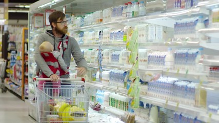 Front view of Caucasian man in glasses carrying baby in sling, talking to child and putting bottle of milk into shopping trolley at supermarket