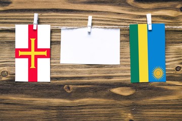 Hanging flags of Guernsey and Rwanda attached to rope with clothes pins with copy space on white note paper on wooden background.Diplomatic relations between countries.