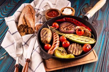 Grilled sausages and vegetables in a pan, towel, mayonnaise, olives and ketchup in wooden bowls, broken buckwheat baguette and cutlery on a blue wooden background.