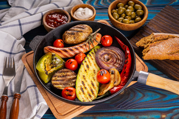 Grilled sausages and vegetables in a pan, towel, mayonnaise, olives and ketchup in wooden bowls, broken buckwheat baguette and cutlery on a blue wooden background.