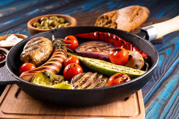 Grilled sausages and vegetables in a pan, towel, mayonnaise, olives and ketchup in wooden bowls, broken buckwheat baguette and cutlery on a blue wooden background.