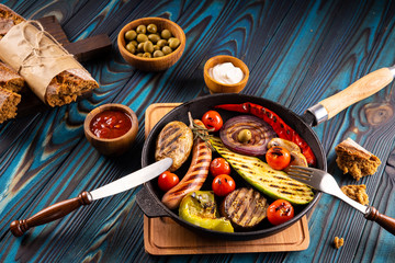 Grilled sausages and vegetables in a pan, towel, mayonnaise, olives and ketchup in wooden bowls, broken buckwheat baguette and cutlery on a blue wooden background.