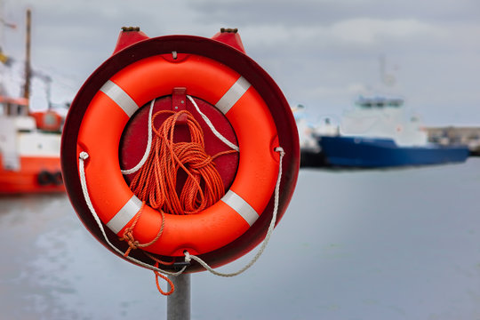 An orange colored lifebuoy in a holding device at the pier in the East German port of Sassnitz. In the background fishing ships out of focus with beautiful bokeh.