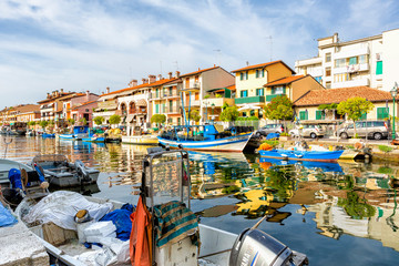 JULY 22, 2019 - GRADO, ITALY - Colored residential buildings and boats in the sunset light are mirror reflected in sea water of the old harbor of Grado, Northeastern Italy - obrazy, fototapety, plakaty