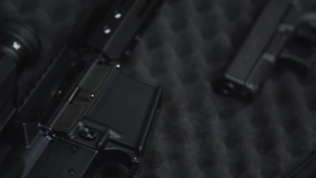 Handheld tracking shot left to right, panning over an arsenal of firearms laid out in an open case containing a sniper rifle, a scoped assault rifle and a 9mm handgun.