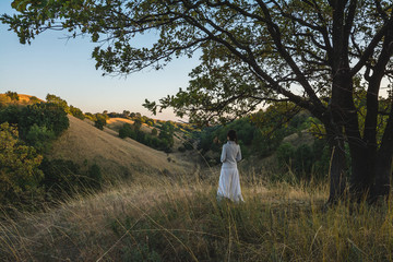 Young woman in white clothing standing under the tree and watching autumn evening sky over the hills of Deliblatska pescara (Deliblato sands) in Serbia