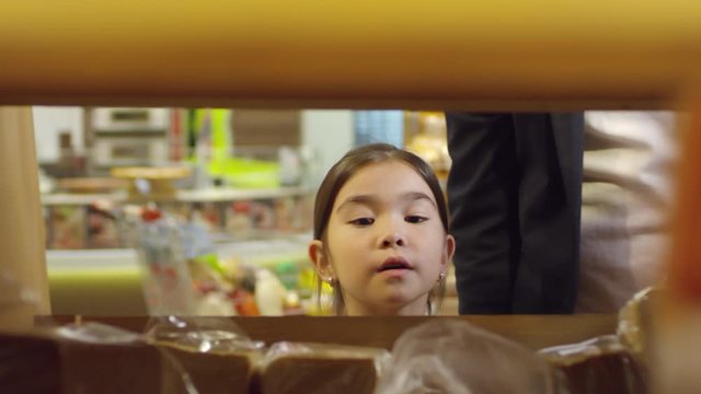 Close up shot of curious little girl walking along supermarket aisle and choosing bread, then taking it from shelf and going away