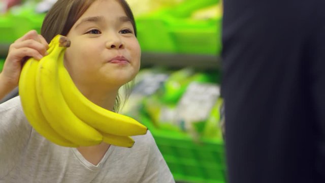 Medium shot of cute Asian girl of primary school age taking bananas at supermarket and showing them to father while shopping together