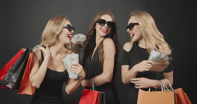 Glamorous women in dark dresses holding paper bags demonstrating dollar banknotes, rich ladies go shopping