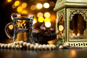 Burning candle in golden lantern with wooden rosary, dates in a bowl and cup of water on bokeh background. Oriental background. Ramadan concept. Selective focus.